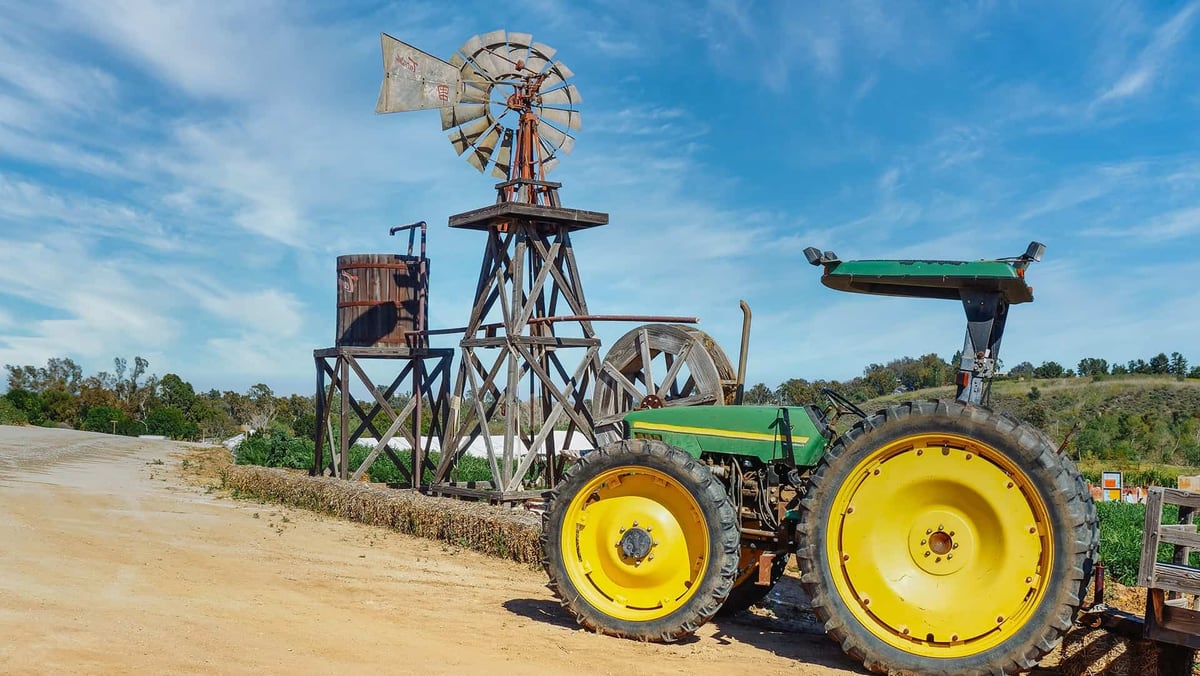A green tractor with yellow wheels parked on a dirt track beside a wooden windmill and water tank, under a blue sky.