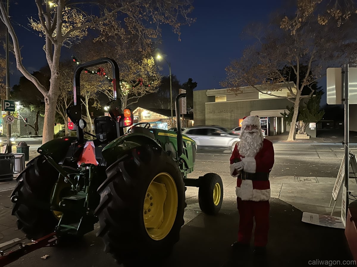 A green tractor parked on a lit street at night, Santa standing beside it.